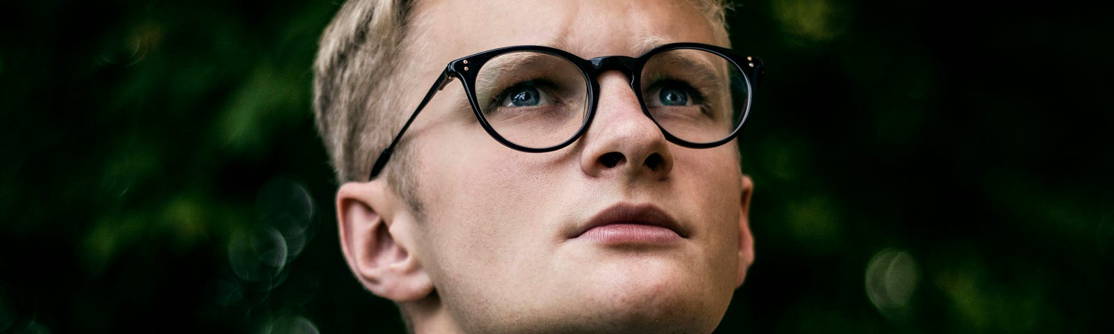 A blonde teenager boy with glasses looks upwards, in deep contemplation