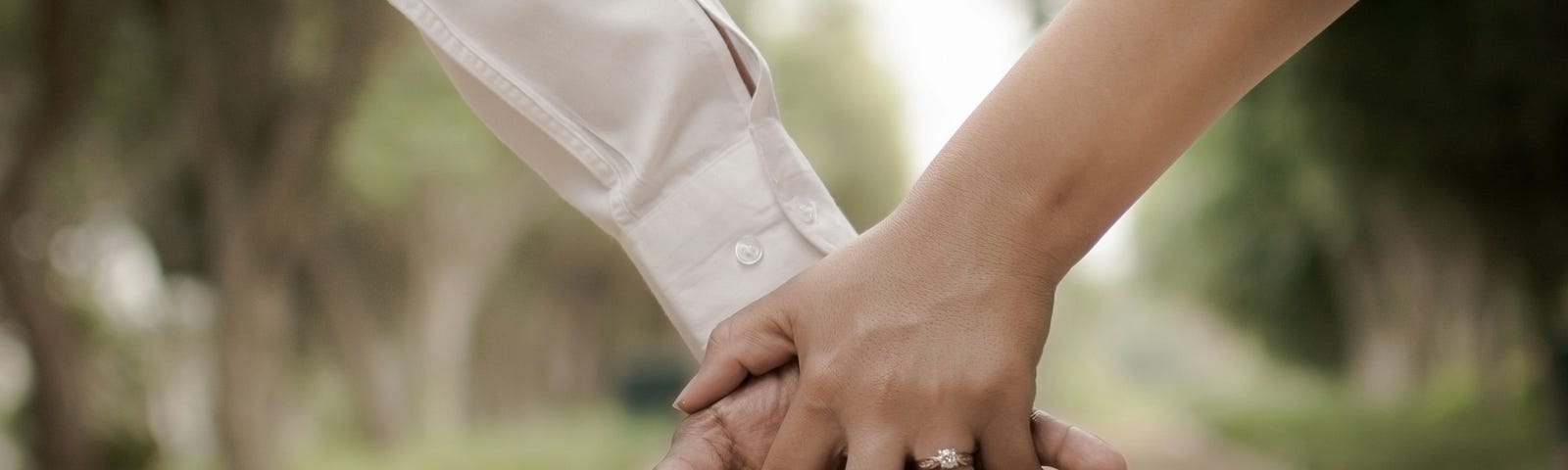 Two hands clasped, one with an engagement ring, and a blurred walkway in the background