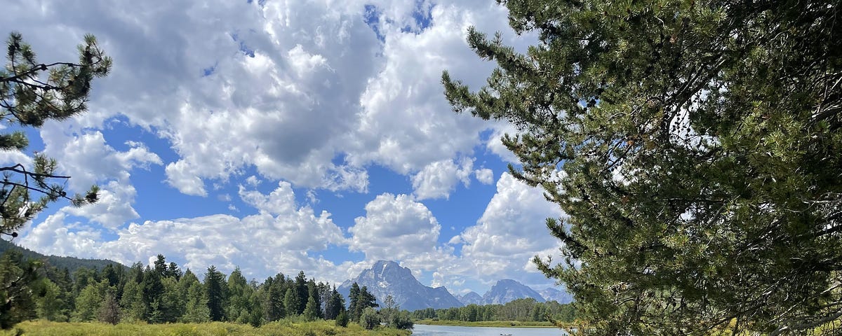 A cloudy sky on a sunny day, with snow-capped mountains in the distance. In the foreground is a lake with evergreens surrounding it.