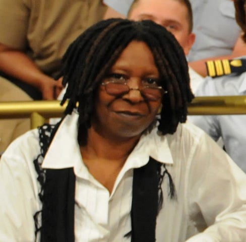 Photo of Whoopi Goldberg with dreadlocks and eyeglasses worn low on her nose. She is wearing a white blouse and a black scarf with minimal fringe. Behind her is a member of the U.S. military.