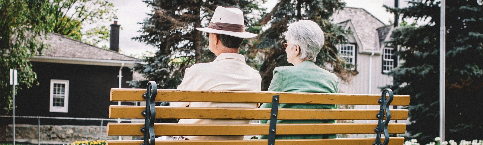 A man of retirement age wearing a Panama hat sits on a slat bench with his female companion peering across a green area covered in green grass and clover within view of homes and a colorful flower bed.