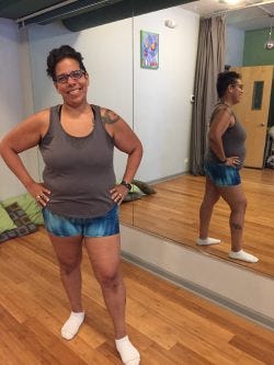 Woman stands facing camera, hands on hips, smiling, wearing short shorts and a tank top. Her full-body reflection in a mirror is also visible.