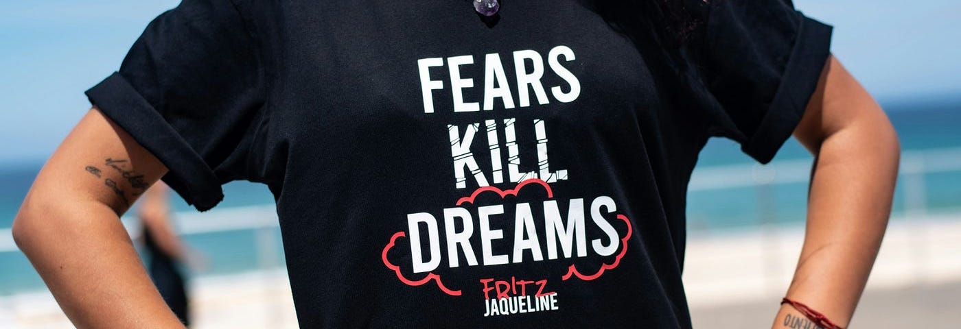 A woman wearing a short sleeve black t-shirt that says Fears Kill Dreams with dreams circled in red. Underneath the slogan is the name Fritz Jacqueline.