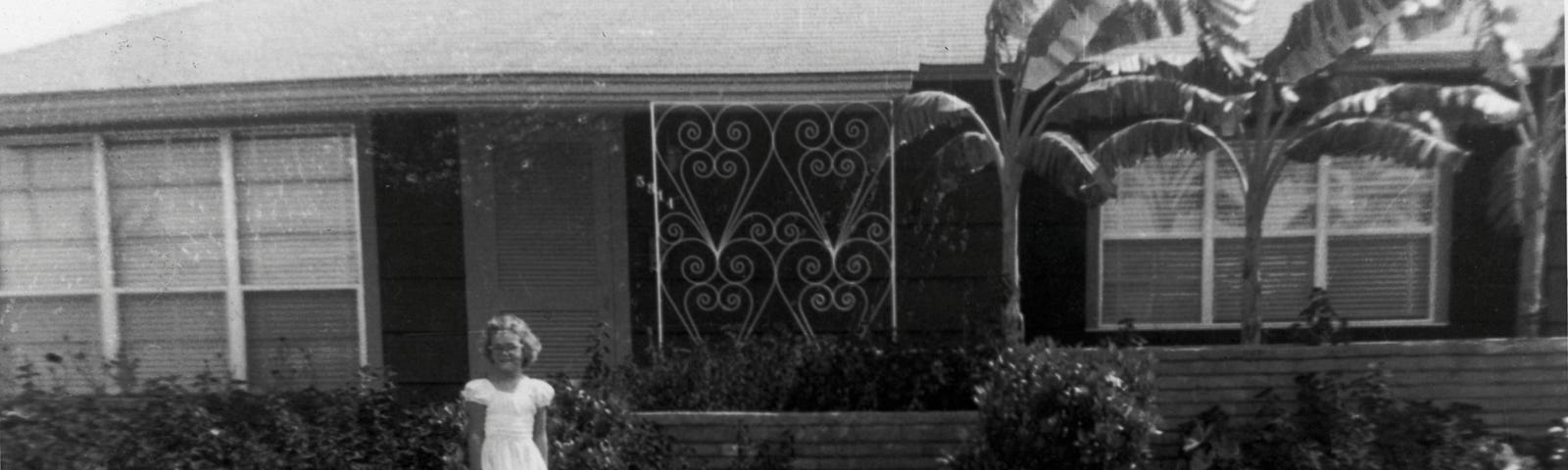 black and white photo of young girl standing in front of a 1950s house in the suburbs-houses and memories