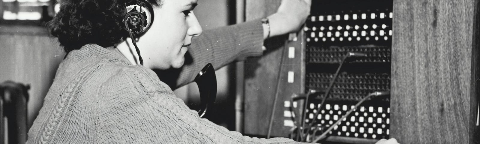 A phone operator circa 1950’s operates a switchboard. She’s wearing a headset and dialing a rotary dial.