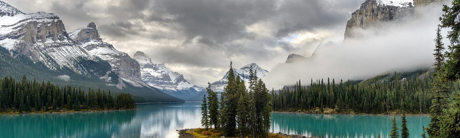 A small stand of spruce trees on a small island on a turquoise lake. There are mountains dusted with snow in the background which are reflected in the lake.