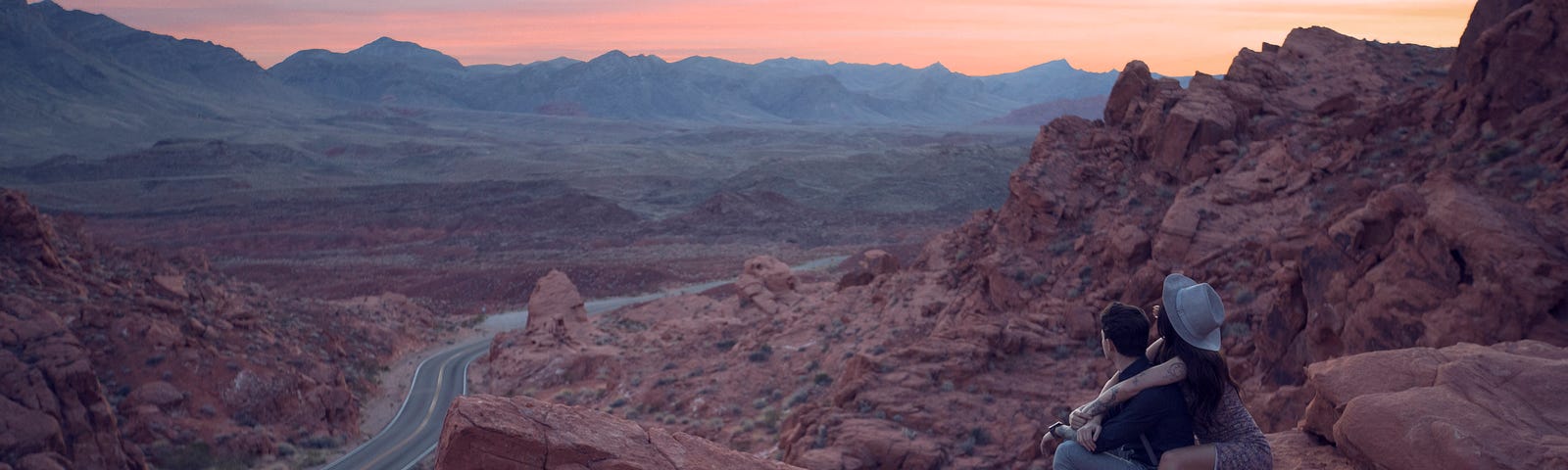 A couple sit together on a red rocky landscape amiring the panoramic view.