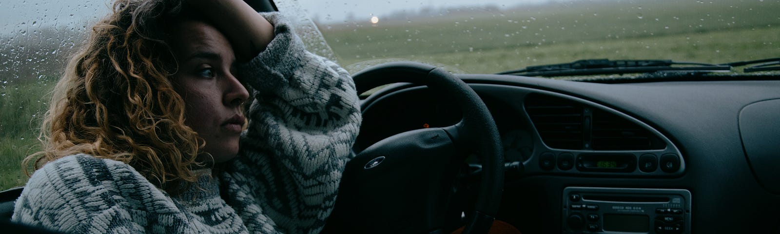 A woman sitting in her car, looking depressed on a dark and rainy day.