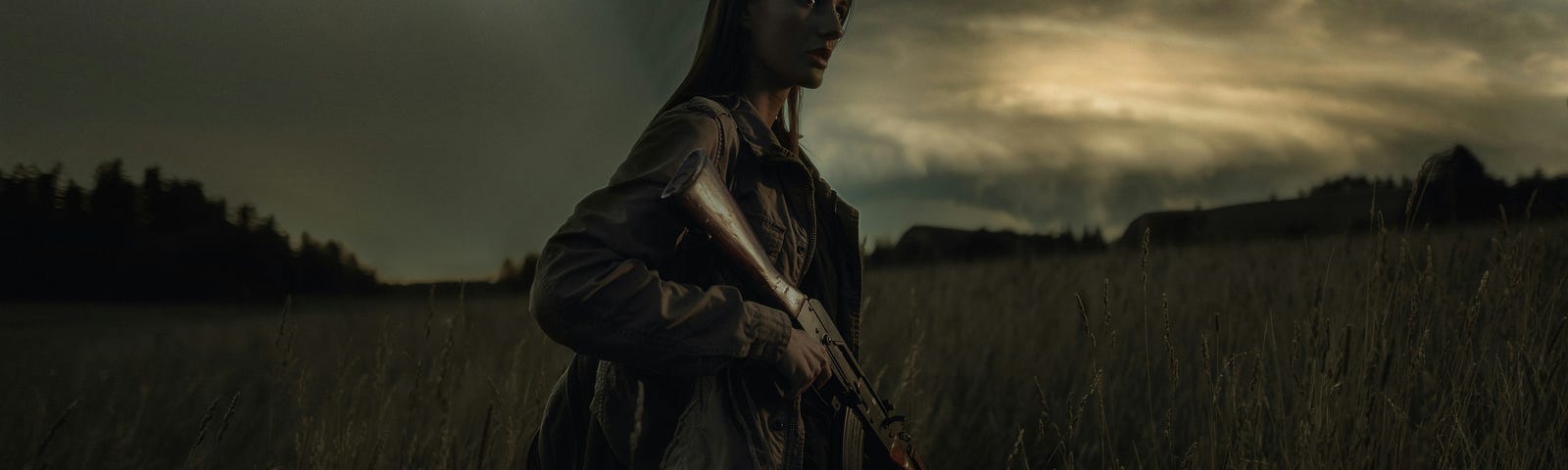 A woman with a rifle on a field of high grass in the evening twilight.