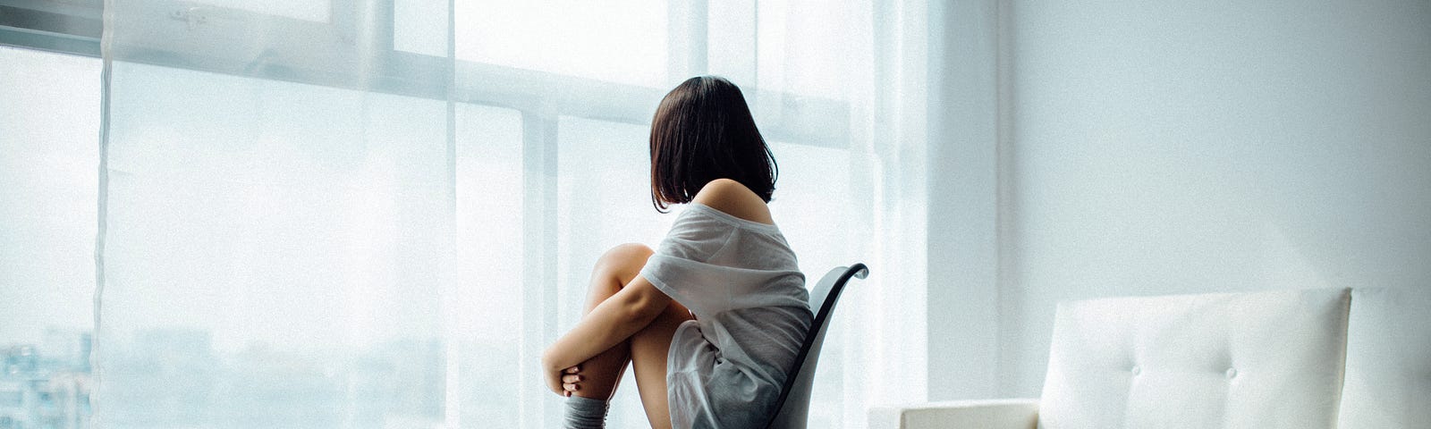 Girl sitting with feet raised facing the window in a cheerless room