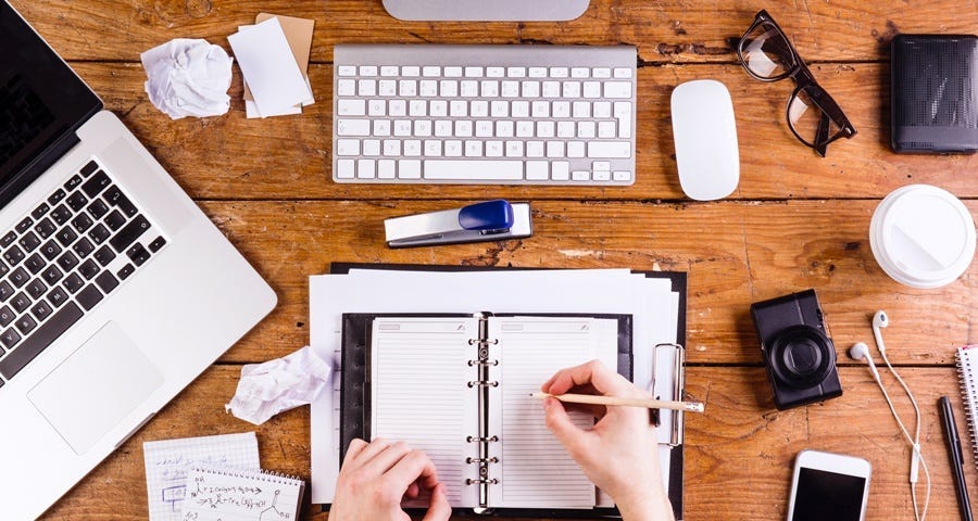 A wooden desk displays many different writer’s tools including a wireless keyboard, mouse, notebook computer, reading glasses, smart phone, pen and paper and notes.