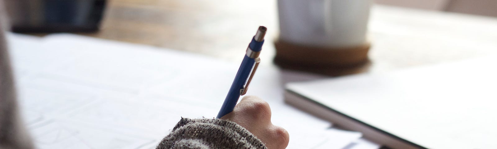 A woman hand writing a letter , a cup on the table and some notebooks lying around