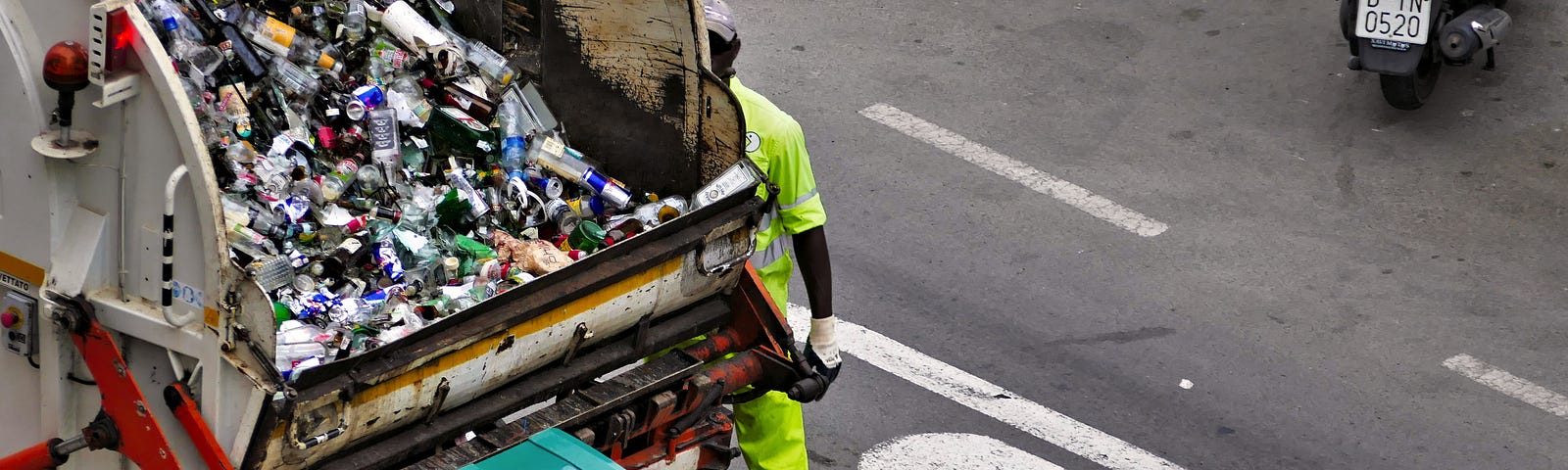 the image is that of the back of a dump truck that is carting away household waste; it is parked in the street and there are two men loading a cart into the back.