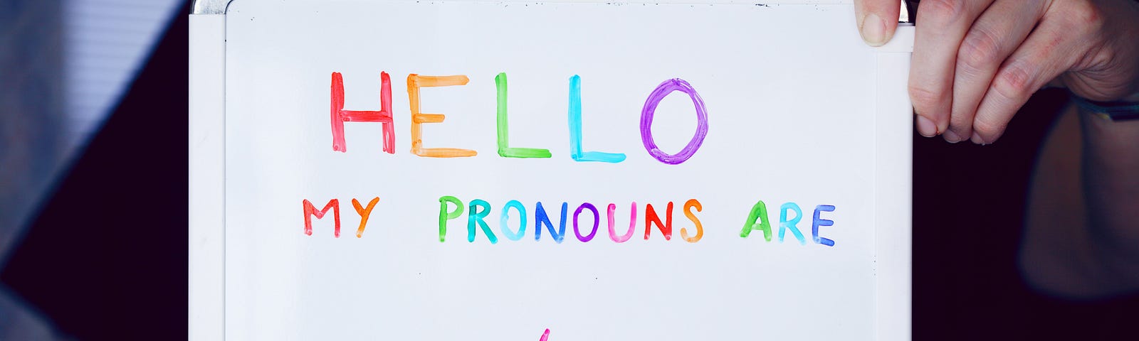 person holding whiteboard sign with rainbow lettering: HELLO MY PRONOUNS ARE with blank underlines and a slash mark to fill in the pronouns