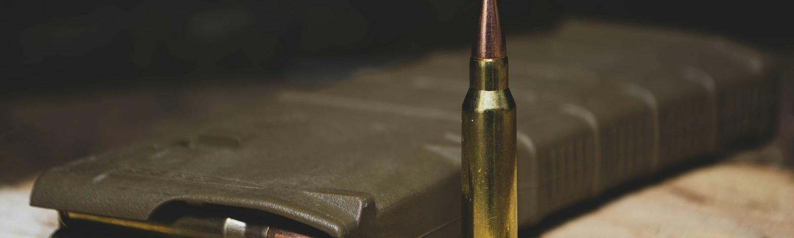 A loaded ammo clip for an automatic rifle laying on aa wooden surface. Next to it are a bullet standing upright and another laying flat on the surface.