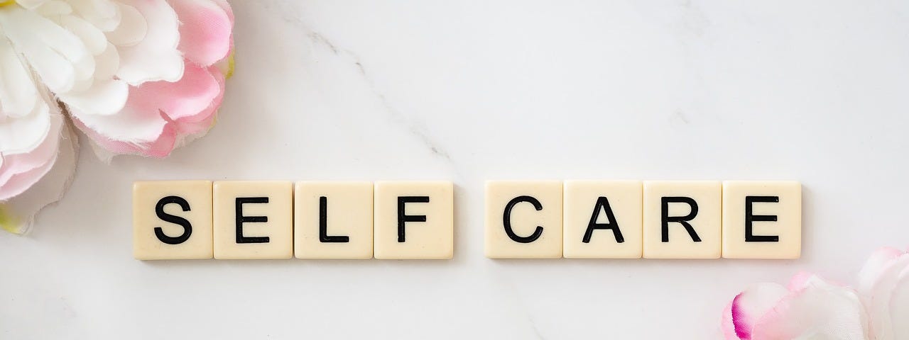 Blocks Of Letters That Spell The Word Self Care In Black Capital Text On A White Background With Some Flowers.