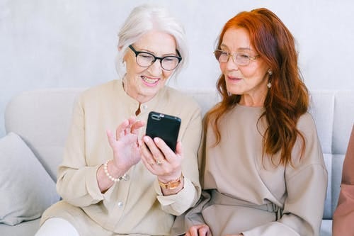 Two women in beige jumpsuits look at a smart phone together, while sitting on a sofa. One woman has white hair and the other woman has red hair. Both are wearing glasses.