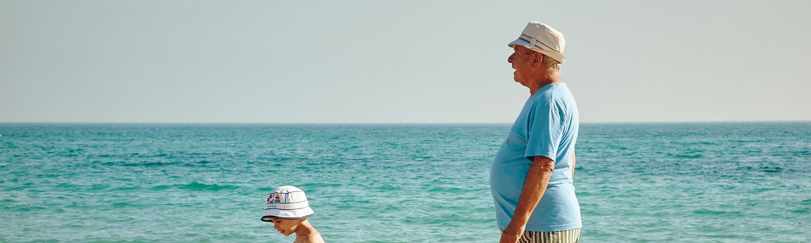 Walking along the beach, with the water in the background, a young child and, presumably, his grandfather. Both are wearing the same kind of sun hat.