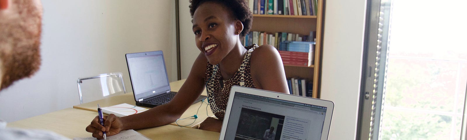 Photo of Palesa Thinta sitting at a desk in an office, writing in a notebook and smiling at Lloyd Belton. He’s sitting across from her and working on a laptop.