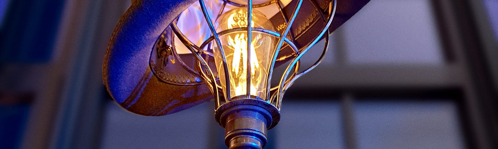 A collection of a metal-caged standing lamp with visible bulb, a hat, and an old style vintage camera box set near the neck of the lamp gives the entire ensemble a semi-human look.