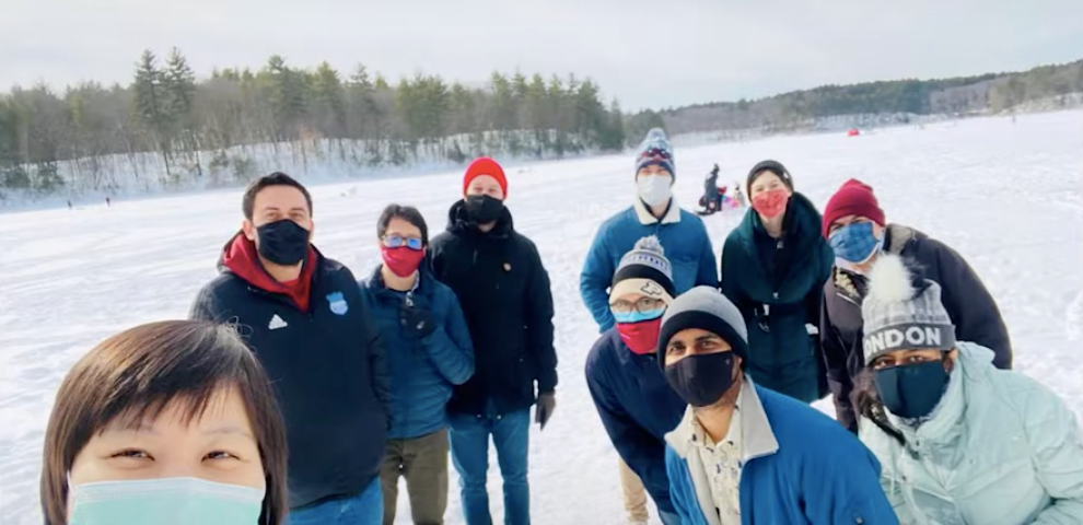 Multiethnic group of men and women dressed in winter wear and face masks, standing on a frozen lake.