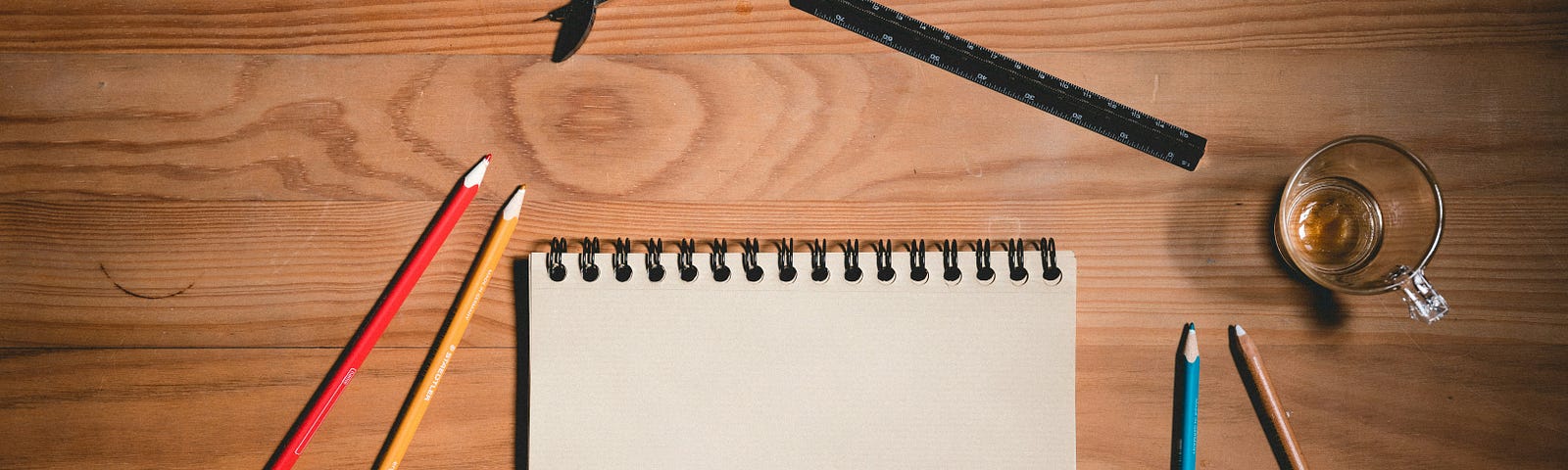 A sketchbook with pencils, pens, and a compass sit on a wooden table. An empty glass mug sits to the right of the notebook.