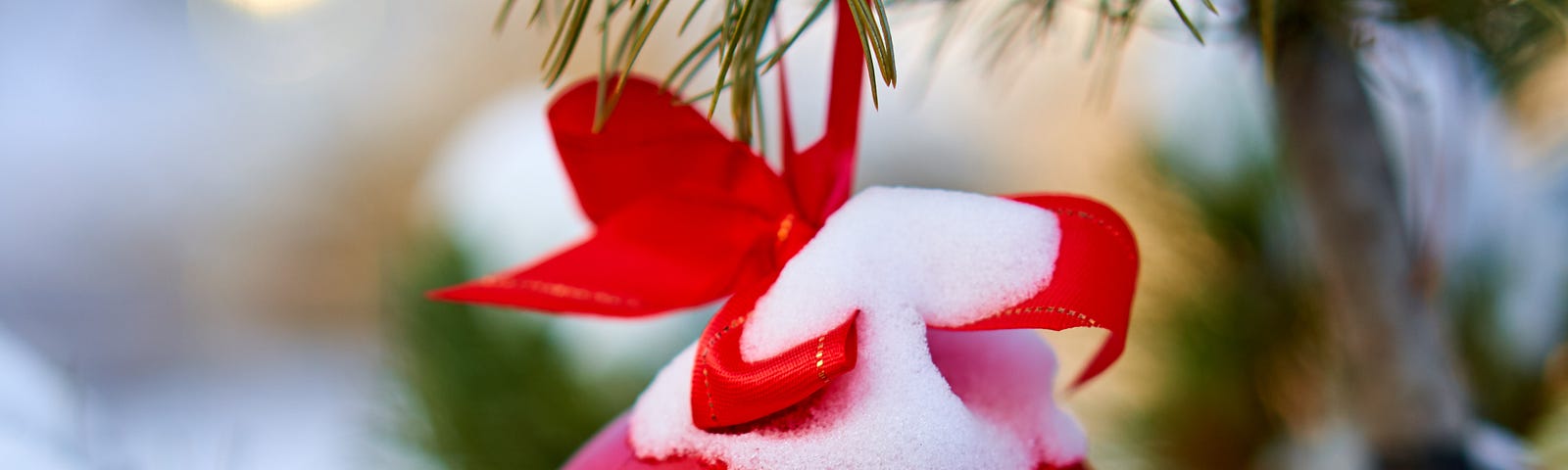 Snow covered red ball ornament hanging from a pine bough by a red ribbon