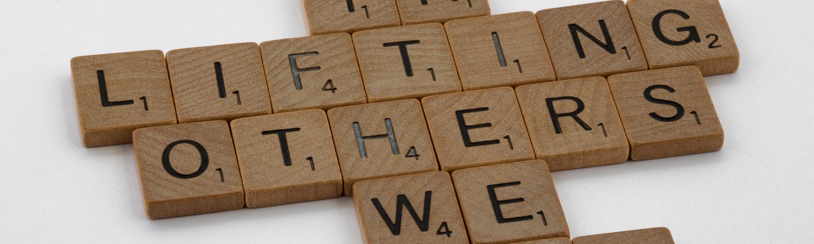 A set of wooden scrabble pieces arranged to read the sentence, “In Lifting Others We Rise”