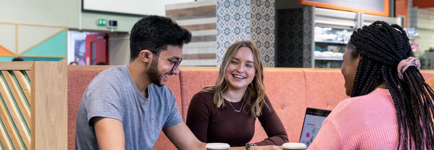 Three students are sat at a cafe table happily chatting to one another while working on a laptop and drinking coffee.