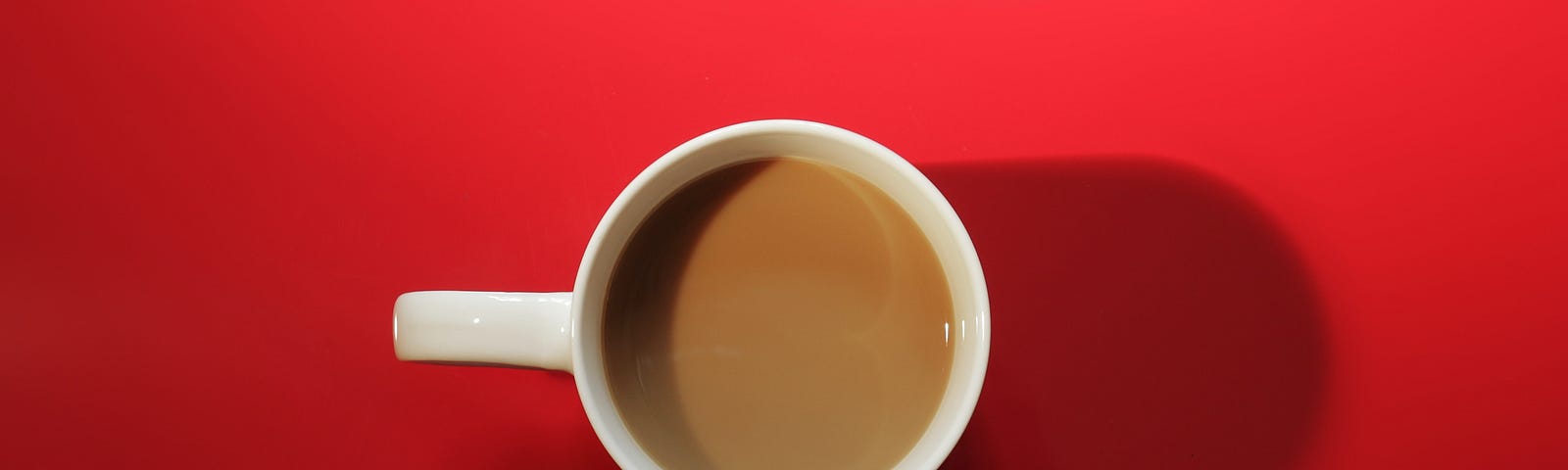 A cup of coffee with cream in a white mug viewed from above casts a shadow on a bright red background.