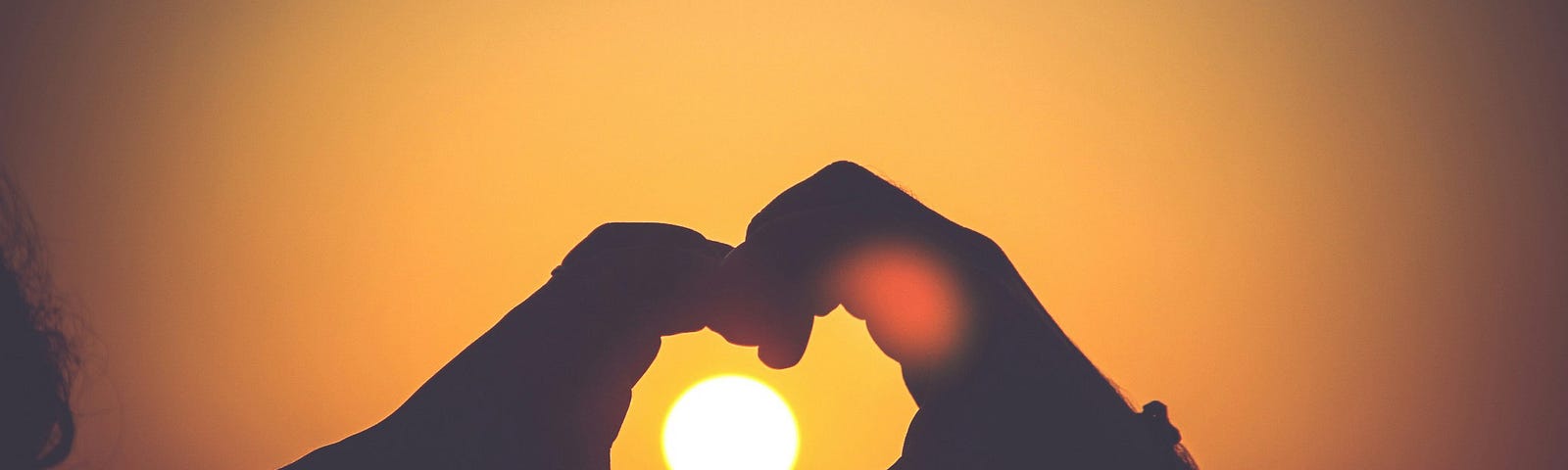 Photograph of two hands forming a heart silhouette, against a golden sky, with the white circle of the setting sun, caught in the heart shape between the hands.