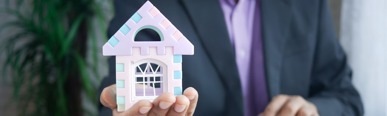 A man in a purple suit jacket sits at a desk using a laptop computer with a house in his hand — the concept of financial management