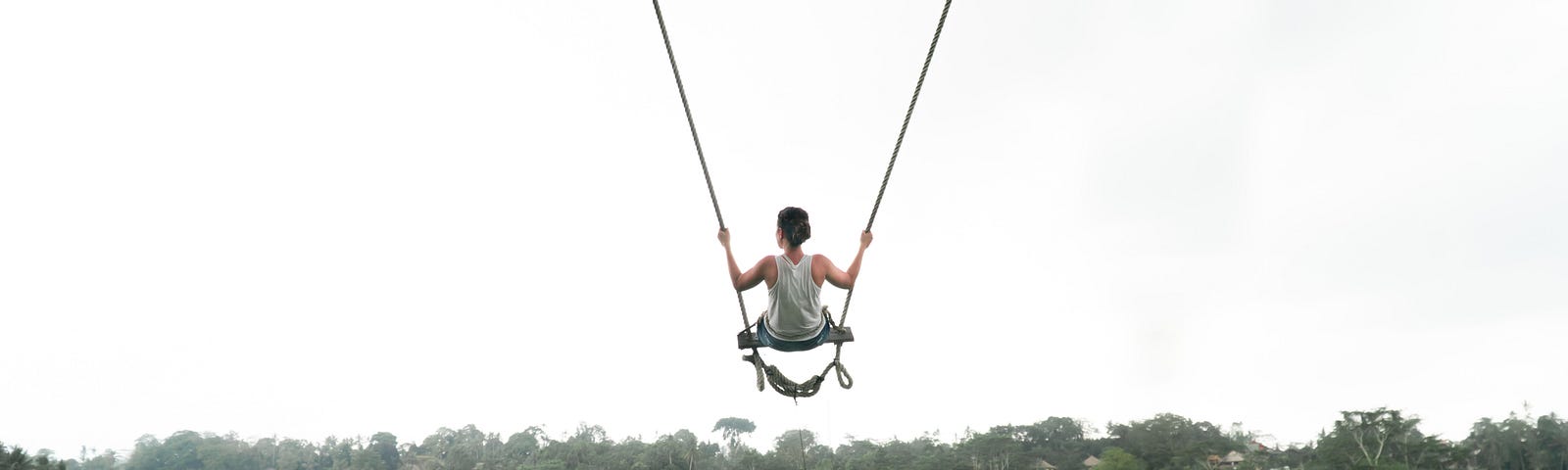A horizontal photo of a woman wearing all white and her long brown hair up in a bun. She is captured swinging forth on a wide wooden swing with very long ropes off the edge of a cliff overlooking the treetops of a great luscious green forest. She is looking to her left as if distracted by wonderment. Itchy Feet: A poem about feeling wanderlust with a pauper’s pockets by Rambling Rose.