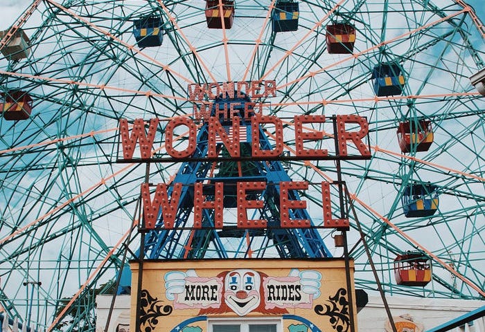 Close up of large, colorful ferris wheel with a yellow sign with a vintage clown face at the entrance that says ‘Thrills and Excitement this way’.