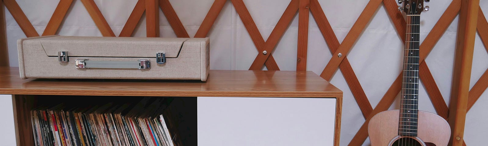 A wood cabinet with white doors has a centre compartment for vinyl records and a portable stereo in a case on top. There is an acoustic guitar leaning against the wall to the right of it.