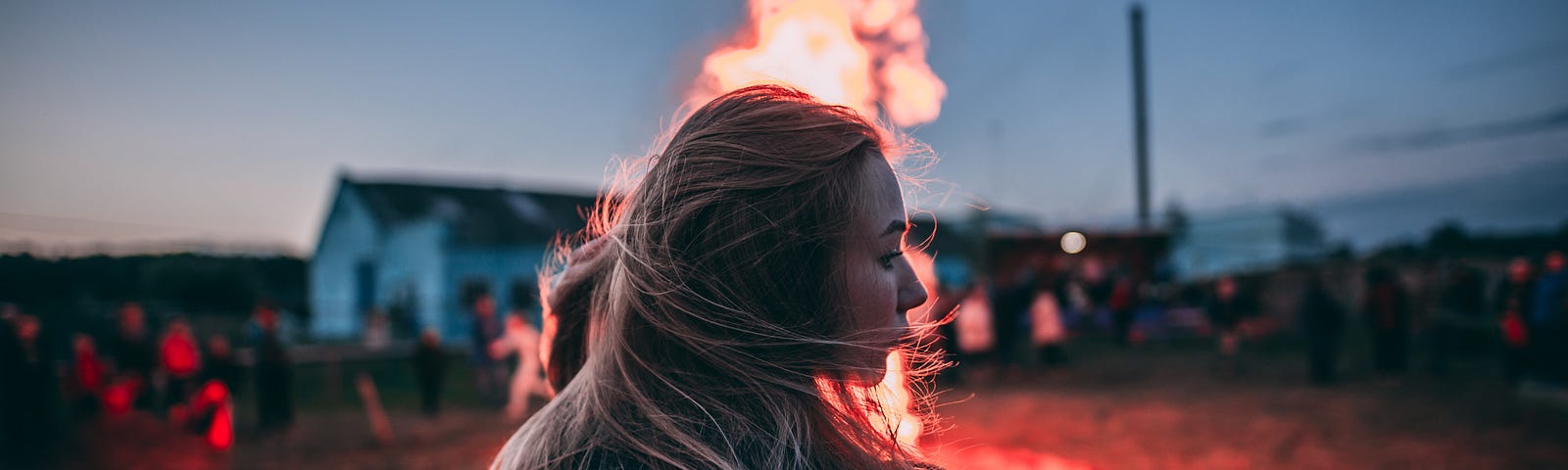 blonde woman with back to the camera standing outside, in front of a bonfire with a house behind it in the far distance