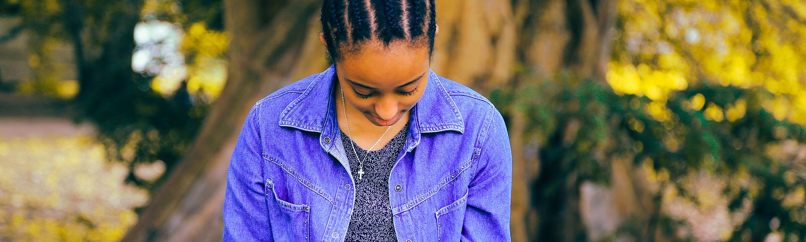 person sitting under a tree, they are wearing blue denim jeans and jacket, they are holding a pen and writing in a notebook