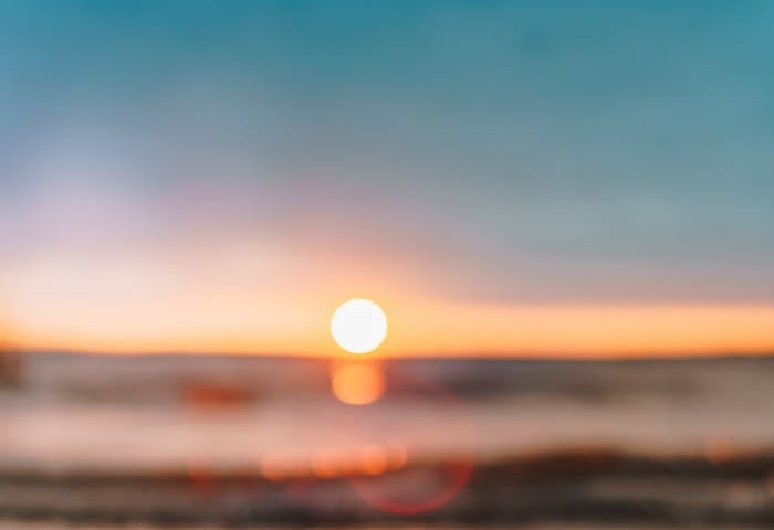 A sunset horizon view on the beach from the sand overlooking a lying glass jar with little string lights inside (that mimic the look of fireflies) and a receding wave. The similar glow of the sunset jar lights and sunset are like bulbs from the same company of lights.