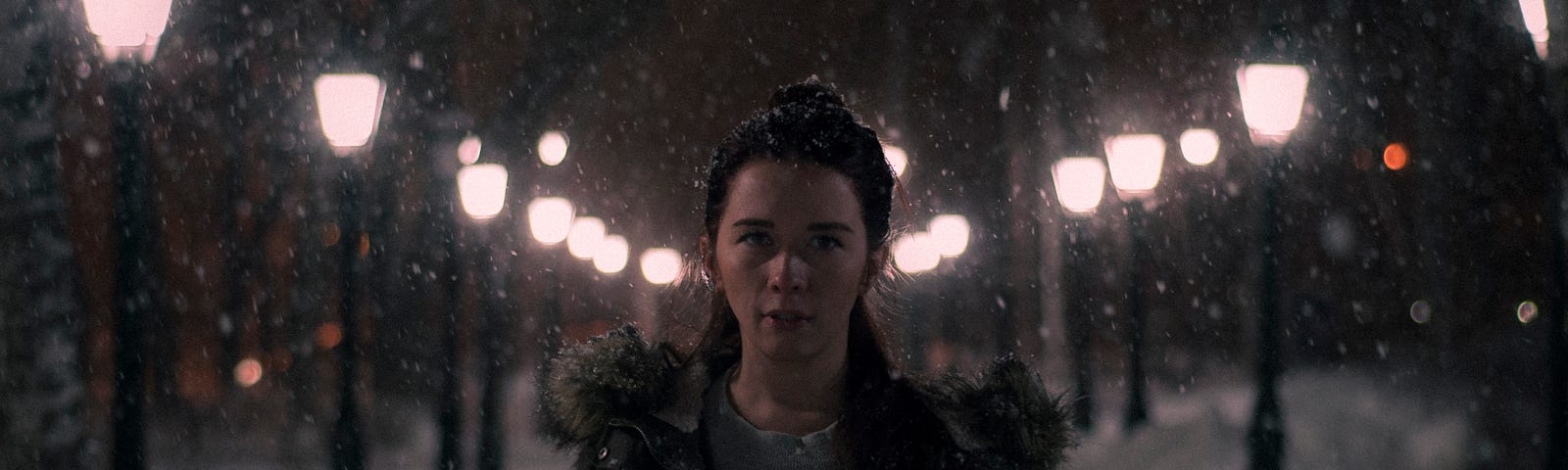 A girl with an open winter’s jacket and fur-lined hood standing at night in front of a lamp-post lit path (all snowed on the path) and looking at the camera. The shadowy look is metaphorical to being the heroine in this story (not glorious or glamorous).