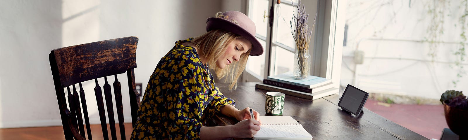 White woman wiht blond hair and a mauve hat writing on a notebook on a wooden desk and chair in a courtyard.