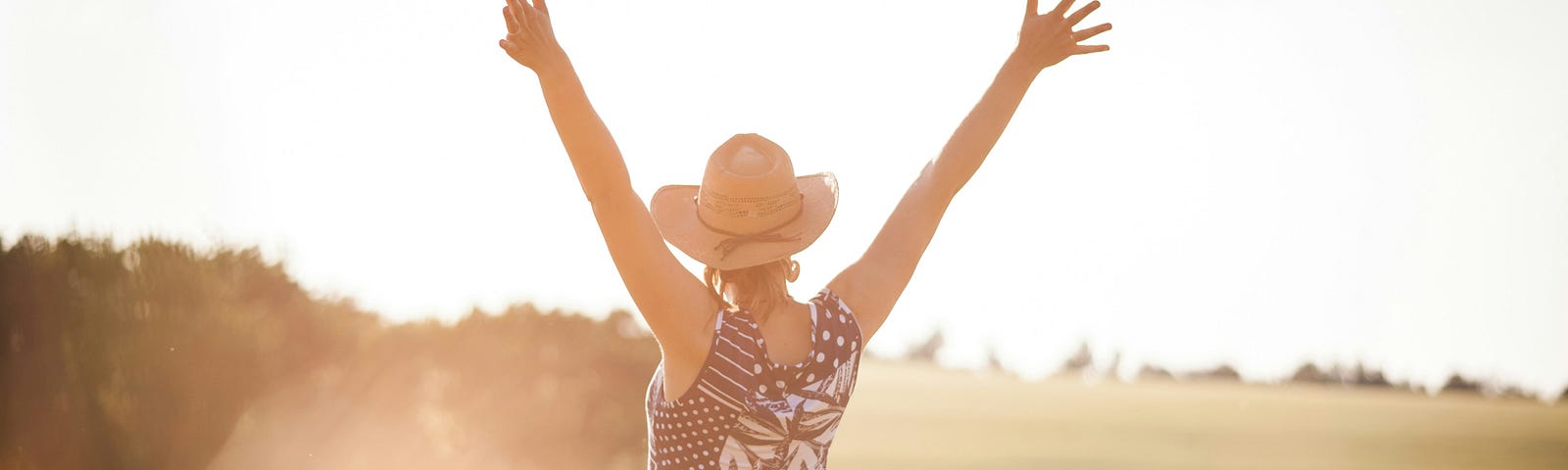 A person wearing a sunhat and dress with arms outstretched upwards in a sunlit field, embodying the feeking of freedom