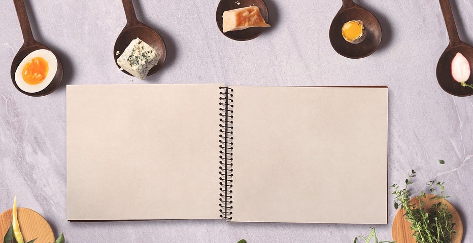 A blank recipe book open, surrounded by wooden spoons holding cheeses, eggs, herbs, garlic, and other ingredients.