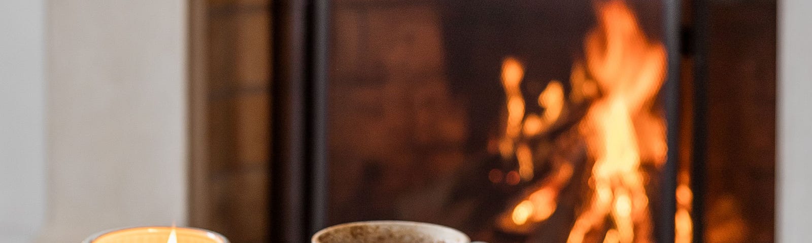 A warm coffee, a glowing candle and a notebook and pen, in front of a roaring fire in a fireplace.