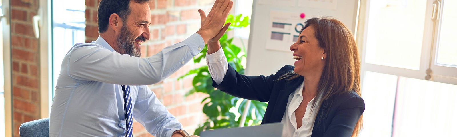 Two business people high-fiving over a desk which holds a laptop, papers, coffee mugs and other business paraphernalia. In the background there are windows letting in light and a whiteboard with graphs and data pinned to it