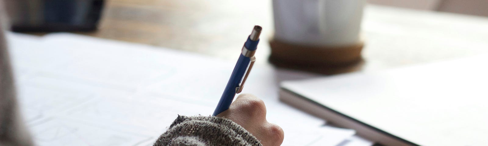 A person writing on paper with a mug and notebook off to the side.