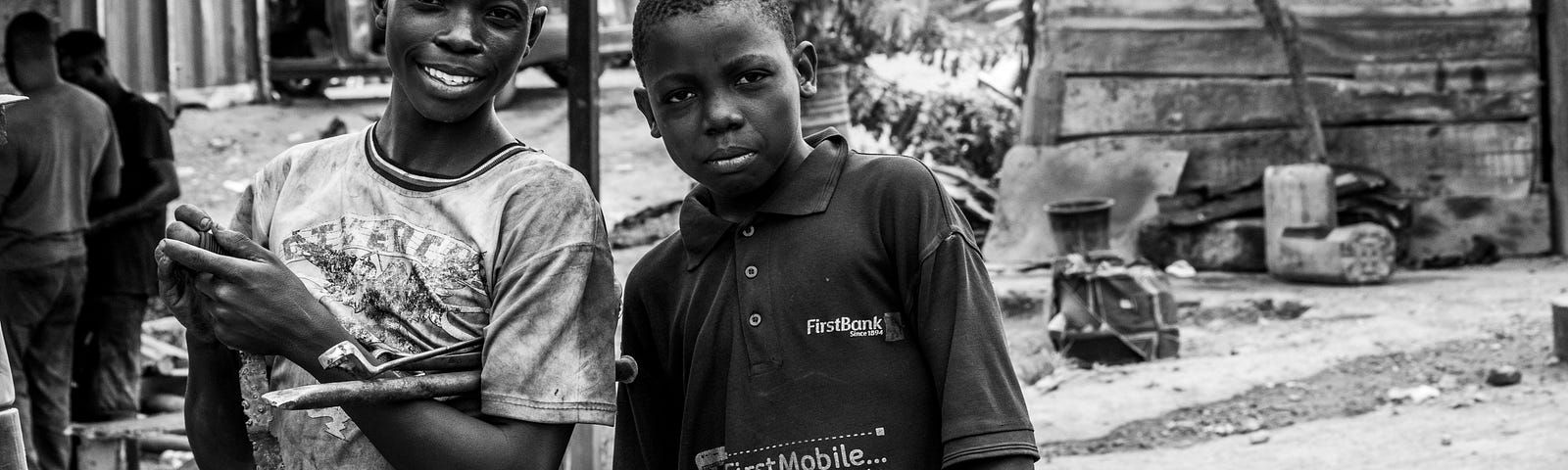 Two teenage boys wearing t-shirts looking into camera. One of them is smiling. There is a wooden shack and people in the background