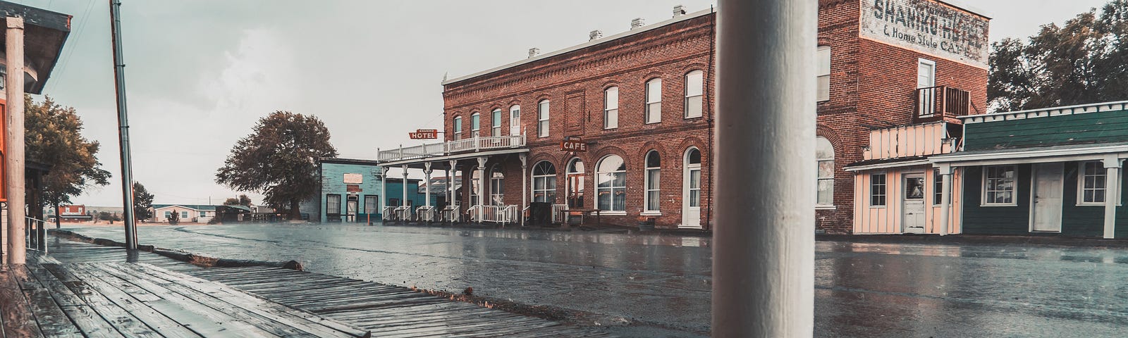 A saloon and shops along the main road of a wild west town. The picture is taken from a porch. The road is wet from recent rain.