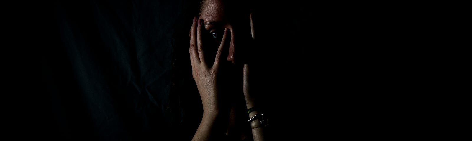 a very dark picture of a woman with her hands on her face. One eye visible, between the fingers of her right hand. the rest of her is in shadows.