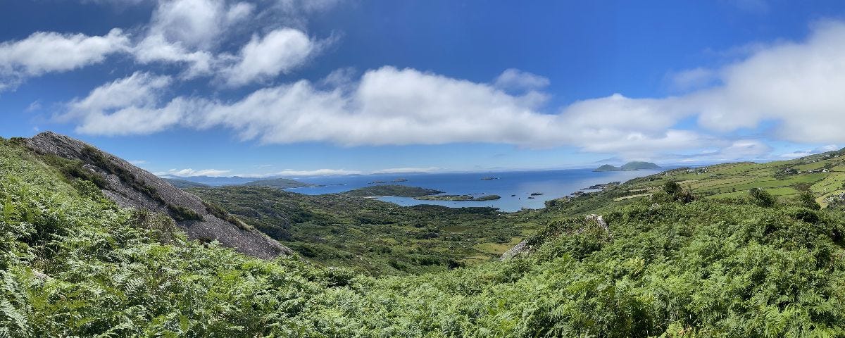 Photo from a hilltop in Derrynane, County Kerry, Ireland, with lush greenery in the foreground and small islands dotting the ocean in the distance. The sky is blue with puffy clouds.