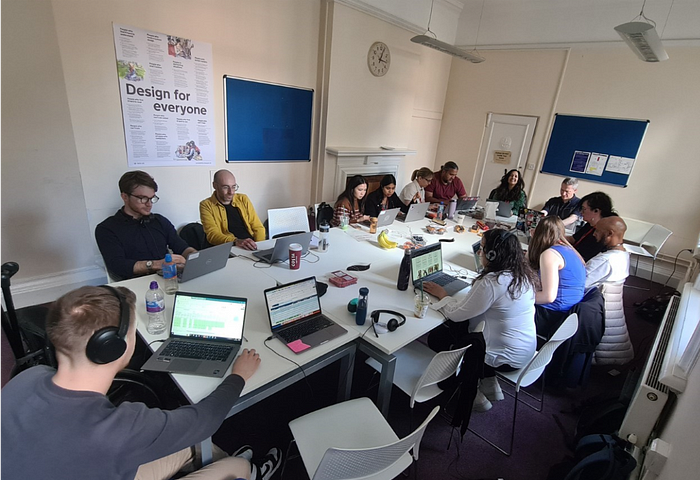 13 people sit around a long table working on laptops. Some have headphones on. A poster on the wall in the background reads ‘Design for everyone’.